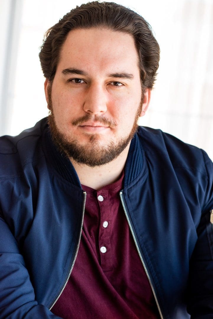 Photo of Greg Harrison looking at the camera stoically, wearing a red shirt and blue jacket.